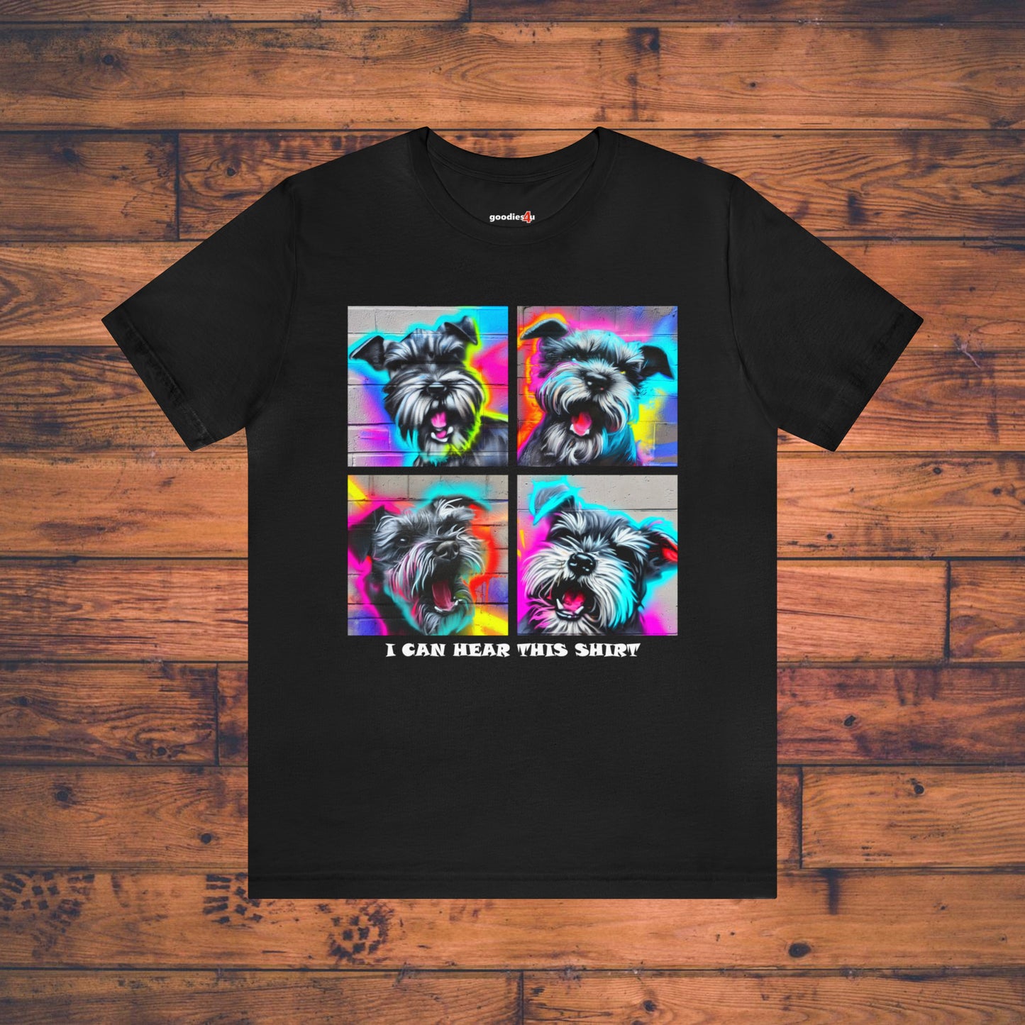 A black t shirt that has four graffiti-style images of barking miniature schnauzers and the text "I Can Hear This Shirt" on a wood floor.