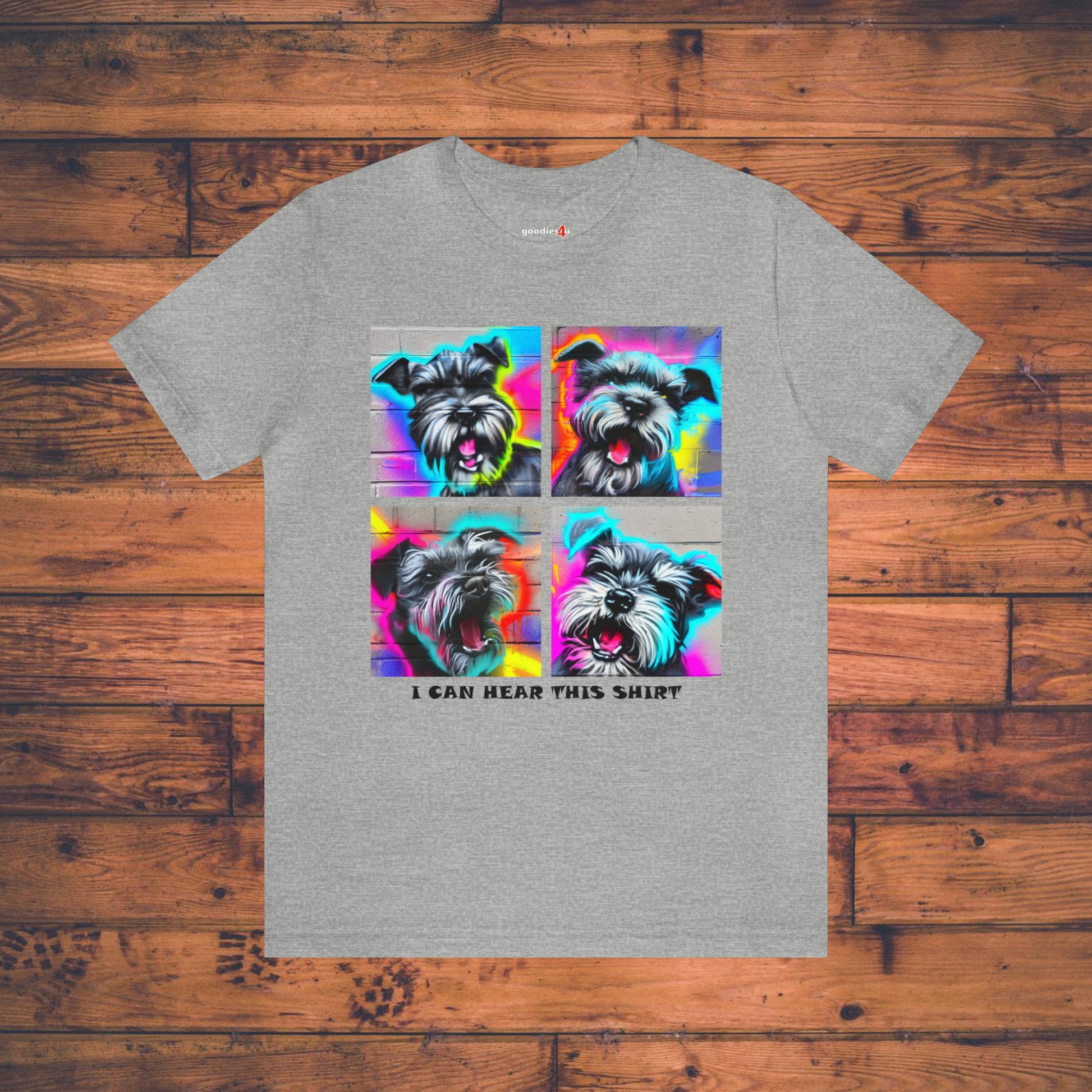 A light gray t shirt that has four graffiti-style images of barking miniature schnauzers and the text "I Can Hear This Shirt" on a wood floor.