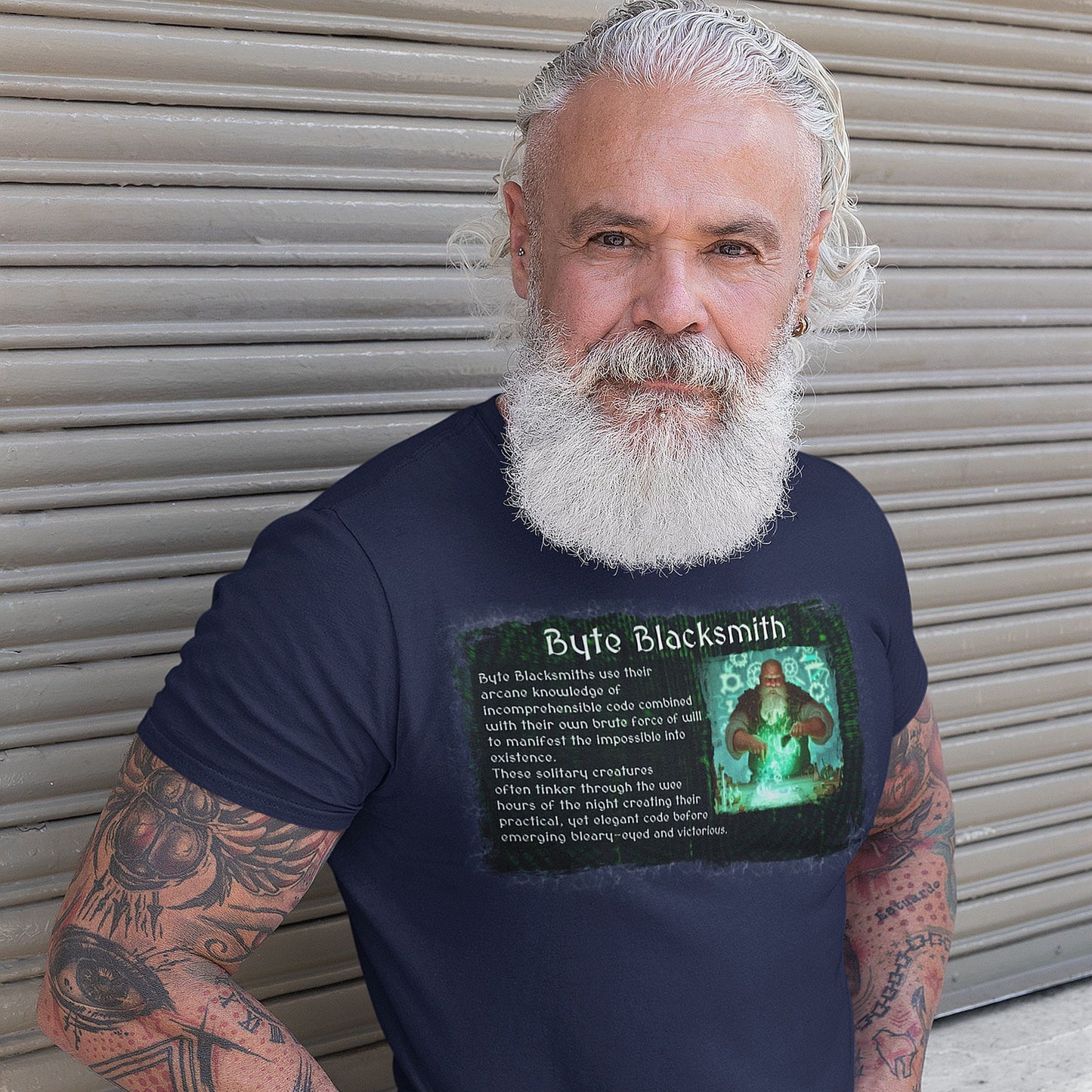 Man wearing heather blue t shirt with the text "Byte Blacksmith" on it. The primary graphic appears weathered and has an image of a bald dwarf with a great white beard in a workshop creating with magic. The RPG-style character description of a software developer as a Byte Blacksmith appears next to the image.